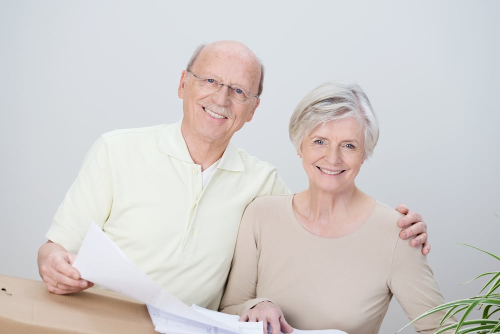 Smiling affectionate senior couple standing with the husbands arm around his wife looking at documents resting on a brown cardboard carton as they plan their move to a new home